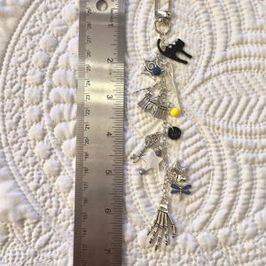 May include: A silver-tone keychain with various charms, including a black cat, stars, a key, and a skeleton hand. The keychain is approximately 18 cm long, as measured by a ruler.