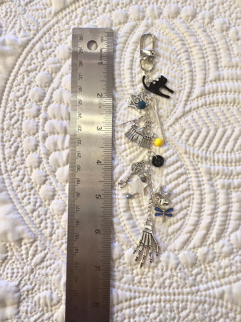 May include: A silver-tone keychain with various charms, including a black cat, stars, a key, and a skeleton hand. The keychain is approximately 18 cm long, as measured by a ruler.