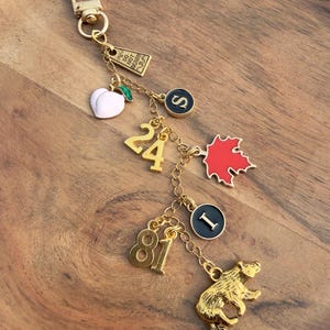 Pode incluir: Chaveiro dourado com vários pingentes. Os pingentes incluem um pêssego branco, uma folha de bordo vermelha, os números 24 e 81, um urso e pingentes circulares com as letras "S" e "I". Um pequeno pingente retangular diz "DO NOT DISTURB".