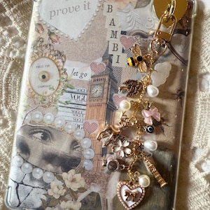 Puede incluir: Funda de teléfono transparente con un diseño de collage que presenta un ciervo, el Big Ben y el texto "prove it". Un dije dorado con una abeja, perlas y un corazón cuelga de la parte superior. La funda tiene una estética vintage.