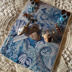 May include: A clear phone case with a blue and white ocean-themed design. The case features two charms with seashells, starfish, and beads. The charms are attached with gold-colored chains and blue clasps.