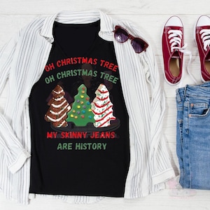 May include: Black V-neck t-shirt with a Christmas tree design and the text "Oh Christmas Tree Oh Christmas Tree My Skinny Jeans Are History". The shirt is paired with a white striped button-down shirt, blue jeans, and red sneakers.