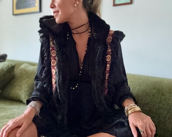 May include: A black dress featuring a sleeveless vest overlay with ruffled edges and a decorative red and gold patterned trim. The outfit includes a thin black choker, layered beaded necklaces, multiple gold-toned bracelets, and black sneakers.