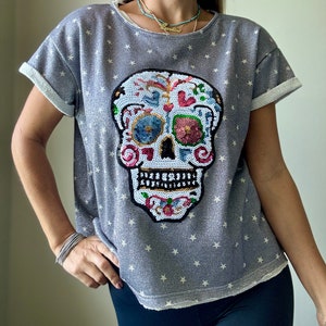 May include: A gray, short-sleeved shirt with a white star pattern and a large, sequined sugar skull design. The skull features colorful floral and heart accents. The sleeves are cuffed with white fabric.
