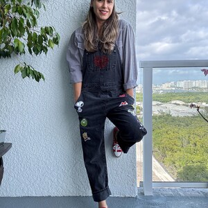 May include: A woman wearing a black overall with a red heart patch and various cartoon character patches. She is wearing a white and black bandana and red converse shoes.