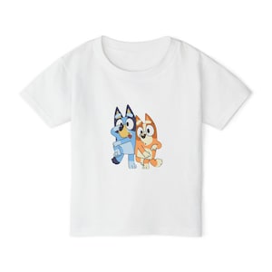 May include: A white short-sleeved t-shirt featuring a central graphic print of two cartoon dogs standing side by side. The dog on the left is blue with dark blue ears, while the dog on the right is orange and cream-coloured with pointed ears.