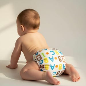 May include: A baby crawling on a white surface, wearing a cloth diaper. The diaper is white with colorful letters and numbers. The baby's skin is fair, and the diaper is the focal point.