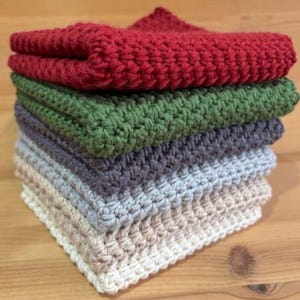 May include: A stack of six square, hand-crocheted washcloths in shades of red, green, grey, light blue, and beige. The washcloths are made from a thick, textured yarn and are neatly stacked. Each cloth is approximately 25 cm square.