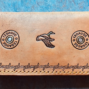 Puede incluir: Cartera de cuero color canela con un emblema de pato y dos emblemas "12 GAUGE". Los bordes están decorados con diseños de alambre de púas. La cartera es rectangular y tiene un borde cosido.