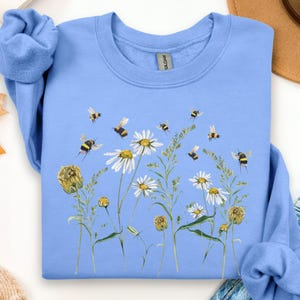 May include: A light blue sweatshirt featuring a floral and bee design. The graphic includes white daisies, yellow wildflowers, and flying bees. The crew neck sweatshirt is made of a soft material.