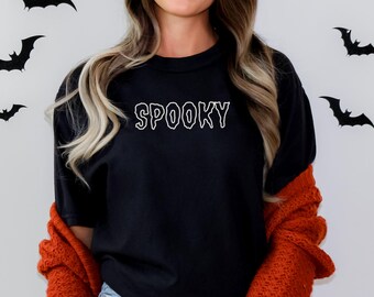 May include: Black t-shirt with the word "SPOOKY" in white, outlined letters. The shirt is paired with an orange sweater. Black bat decorations are on the white background.