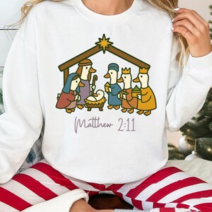 Puede incluir: Sudadera blanca con una escena de la Natividad con patos de dibujos animados. La ilustración incluye una estrella, un establo y el texto "Matthew 2:11". La sudadera la lleva una persona.