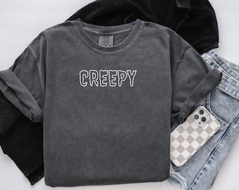 May include: A dark gray, crew neck t-shirt with the word "CREEPY" in white, outlined letters. The shirt is folded, with a black jacket and blue jeans visible in the background. A phone with a checkered case rests on the jeans.