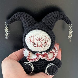 Peut inclure: Une petite poupée crochetée à la main avec une capuche de bouffon noire ornée de breloques en forme d'étoile argentée. Elle possède un visage blanc aux traits brodés en rouge, une collerette blanche à bordure rouge et des membres noirs.