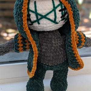Peut inclure: Une poupée en crochet avec une tête ronde blanche ornée de motifs faciaux cousus en vert foncé et noir. Elle porte une cape à capuche vert foncé avec une bordure orange, un torse gris foncé et des jambes vert foncé avec des manches assorties.
