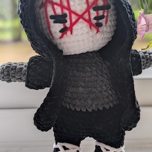 Peut inclure: Une poupée crochetée à la main avec une tête blanche ornée de marques faciales rouges brodées et d'yeux noirs. Elle porte une cape à capuche noire sur un haut gris foncé texturé, un pantalon noir et des chaussures noires à lacets blancs croisés.