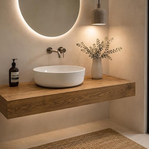 Könnte beinhalten: Ein weißes Keramik-Aufsatzwaschbecken steht auf einem dicken, schwebenden Waschtisch aus Holz. Ein wandmontierter Wasserhahn aus dunklem, mattem Metall befindet sich darüber. Ein runder, hinterleuchteter Spiegel, eine Pendelleuchte und eine Seifenflasche.