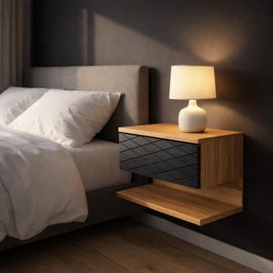 Op de afbeelding: Een moderne zwevende nachtkast met een zwarte diamantvormige lade en een onderste plank. Het nachtkastje is gemaakt van hout en wordt verlicht door een witte lamp met een beige lampenkap. Het bed heeft wit beddengoed.
