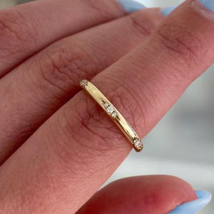 Puede incluir: Un anillo de oro con pequeñas piedras preciosas transparentes engastadas a lo largo de la banda. El anillo se lleva en un dedo con esmalte de uñas azul claro. El anillo parece ser una joya delicada.