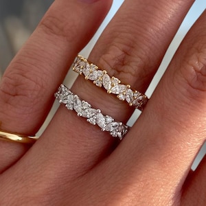Peut inclure: Une main présente trois bagues : une bague en or avec de petits diamants, une bague en or avec des pierres en forme de marquise et une bague en argent avec des pierres en forme de marquise. Les ongles sont peints en bleu clair.