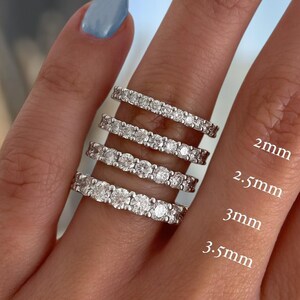May include: A hand displays four silver eternity rings with clear gemstones. The rings are labeled with sizes: 2mm, 2.5mm, 3mm, and 3.5mm. The rings are set against a background of a hand with light blue nail polish.