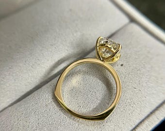 May include: A gold ring with a large, clear gemstone. The ring has a simple band and a four-prong setting that holds the gemstone securely. The ring is displayed on a gray, textured surface.