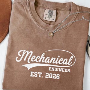 May include: A brown Comfort Colors t-shirt with the words "Mechanical Engineer EST. 2026" in white script. A silver necklace with a small pendant rests on the shirt. A brown hat is partially visible in the upper left corner.