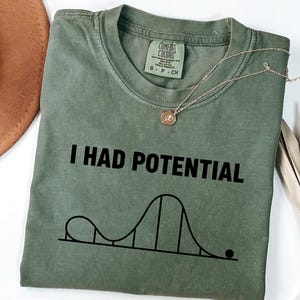 Puede incluir: Camiseta verde oliva con el texto "I HAD POTENTIAL" y un gráfico de montaña rusa. Un collar dorado con un colgante descansa sobre la camiseta. Un sombrero marrón y una planta seca están en el fondo.