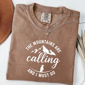 Może przedstawiać: Brązowa koszulka Comfort Colors z białym napisem "THE MOUNTAINS ARE calling AND I MUST GO". Projekt zawiera pasmo górskie i pingwina. Srebrny naszyjnik z wisiorkiem jest ułożony na koszulce.