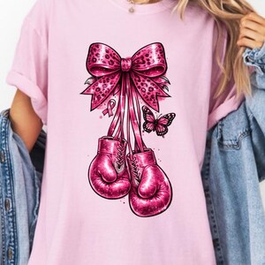 May include: A light pink t-shirt featuring a graphic print of two bright pink boxing gloves tied together with a large leopard-print bow. A small pink ribbon and a butterfly are positioned near the bow. The text ORVIX is printed in black at the bottom.