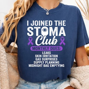 May include: A navy blue short-sleeve t-shirt featuring white text that reads I JOINED THE STOMA CLUB, with two purple ribbons and a list of monthly dues including leaks, skin irritation, gas surprises, supply planning, and midnight bag emptying.