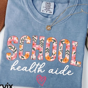 May include: A blue Comfort Colors t-shirt with the words "SCHOOL health aide" in a floral design. A gold necklace and a pink heart graphic are also visible. The shirt is a size small.