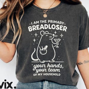 Pode incluir: Uma t-shirt cinzento-carvão de manga curta com um gráfico branco de um rato a segurar um copo de martini e um pedaço de pão. O texto diz: I AM THE PRIMARY BREADLOSER YOUR HANDS, YOUR TEAM OF MY HOUSEHOLD. A t-shirt tem gola redonda.