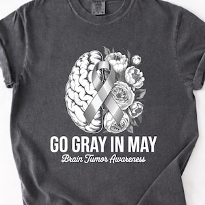 Puede incluir: Camiseta gris oscuro con un diseño que presenta un cerebro, una cinta gris y flores. El texto "GO GRAY IN MAY" y "Brain Tumor Awareness" está impreso debajo. La camiseta es de estilo unisex.