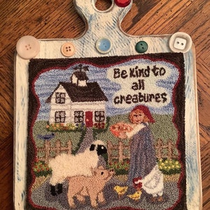 Op de afbeelding: Een decoratieve houten snijplank met een wit en blauw geschilderde handgreep. De plank heeft een gestikt ontwerp met een boerderijscène, inclusief een huis, schapen, een varken en de tekst "Be kind to all creatures". Kleurrijke knopen sieren de randen.