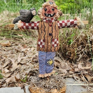 May include: A handmade scarecrow figurine with a brown and red checked shirt, blue jeans, and a bird perched on its shoulder. The scarecrow is standing on a wooden base with brown straw.