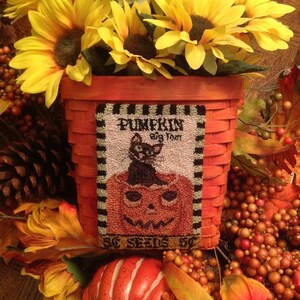Op de afbeelding: Een rode en witte geweven mand gevuld met zonnebloemen en herfstbladeren. De mand heeft een label met de tekst "Pumpkin Big Toot 5¢ Seeds 5¢".