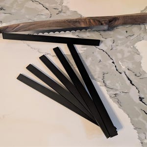 May include: Six black metal strips arranged in a fan shape on a white countertop. The strips are approximately 12 inches long and 1 inch wide.