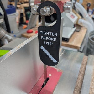 May include: A black warning tag attached to a band saw blade that reads "Tighten Before Use!" The tag has a sawtooth design at the bottom.