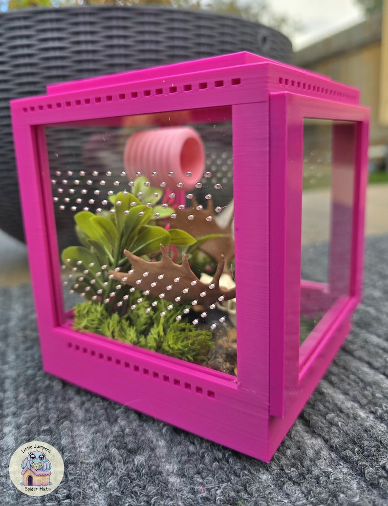 May include: A bright pink terrarium with clear glass panels. Inside, there's a pink tube, artificial plants, and a brown decorative piece. The terrarium is designed with small ventilation holes and is likely for small pets.