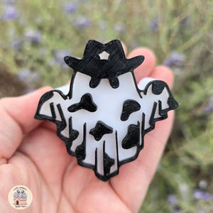 May include: A white and black ghost-shaped pin with a cowboy hat. The ghost has black cow spots and a black outline. The cowboy hat has a star on the front. The pin is held in a hand.