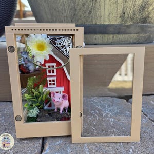 May include: A miniature farm scene inside a rectangular frame. The scene includes a red barn, a pink pig, a gray mouse, and faux greenery. A white flower and a spider web are also visible. The frame is beige, and a separate frame piece is next to it.