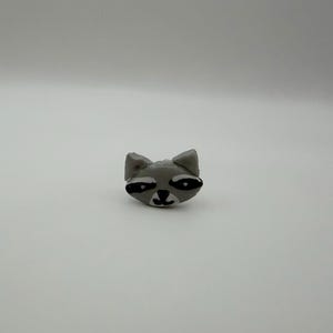 May include: A small, sculpted raccoon head figurine featuring a light gray face with prominent black eye patches, a black nose, and a small black mouth. The raccoon has two pointed ears and a smooth, rounded shape, presented in a neutral setting.