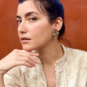 May include: A woman wearing a gold-coloured vintage earring. The earring has an ornate design with intricate details. The woman is wearing a cream-coloured lace blouse. The background is a solid orange colour.