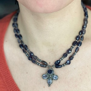 May include: A two-strand necklace featuring dark blue oval beads and a silver-toned pendant. The pendant has a four-petal design with blue and light blue gemstones. The necklace is worn against an orange top.