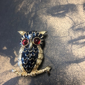 Op de afbeelding: Een uilenbroche van goudkleurig metaal met donkerblauw emaille op de vleugels en borst, voorzien van twee opvallende ronde rode glazen ogen. De uil zit op een gestructureerde goudkleurige tak en vertoont gedetailleerde verenpatronen.