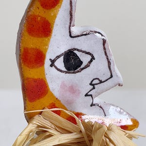 May include: A ceramic face sculpture in a crescent moon shape. The moon features orange and red spots, a black eye, and a pink cheek. The sculpture is decorated with raffia and has a green and white base.