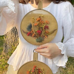 May include: Two round wall hangings with dried flowers. Each features a burlap background, a gold-colored metal frame, and a handle. The flowers are in shades of orange, red, and green, creating a natural, rustic aesthetic.