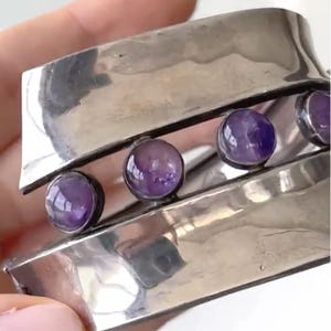 Peut inclure: Un large bracelet manchette en métal argenté poli avec une ouverture horizontale centrale. Trois cabochons ronds et lisses en améthyste violette sont sertis dans des chatons en métal foncé au sein de cette ouverture, répartis uniformément.