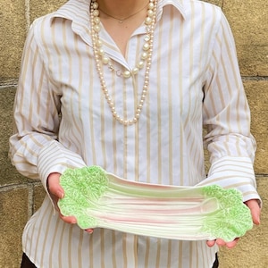 May include: A ceramic serving platter in the shape of a celery stalk, with green, pink, and white hues. The platter is held by a person wearing a striped shirt and a pearl necklace.